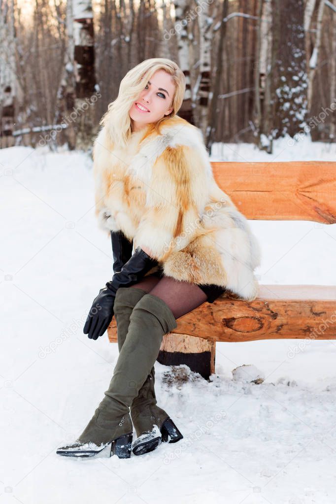 Blonde in fur coat, boots poses on wooden bench outdoor at winter day in park