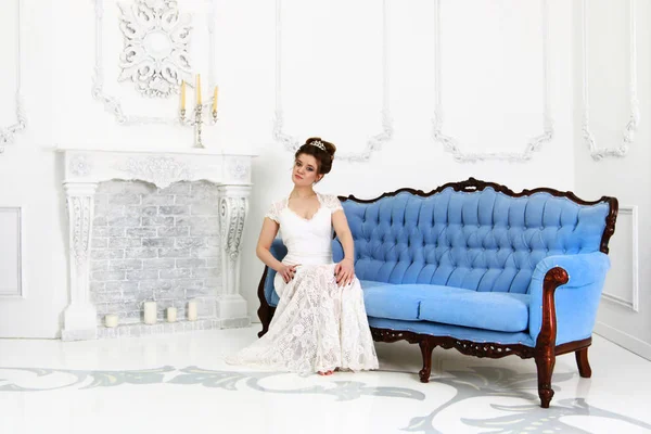 漂亮的年轻新娘穿着白色礼服坐在巴洛克式的沙发在工作室与壁炉 — 图库照片