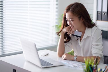 Asyalı çalışan kadınlar internetten alışveriş yapıyor. Kredi kartı tutuyor ve dizüstü bilgisayar kullanıyor..