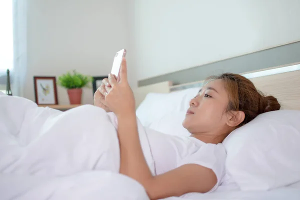 Asian woman is playing a smartphone on a bed in a room.
