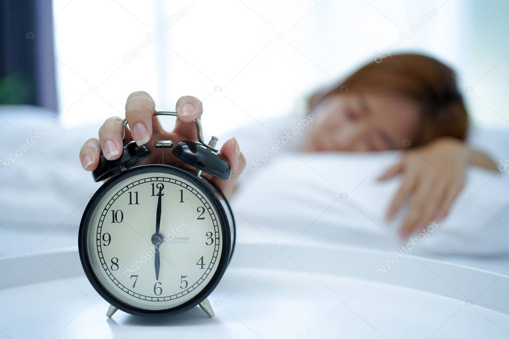 Young Asian woman sleeping in bed, hand pressed the snooze button on clear alarm clock in the morning. Relaxing concept on the weekend
