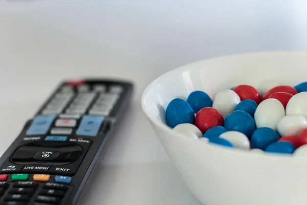 White bowl with red, white and blue candy dragee and tv remote on white table