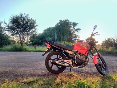 Kırmızı motosiklet manzaralı park yeri, asfalt kaldırımda duruyor ağaçların arka planına ve gökyüzüne karşı gün batımında araçta parlıyor.