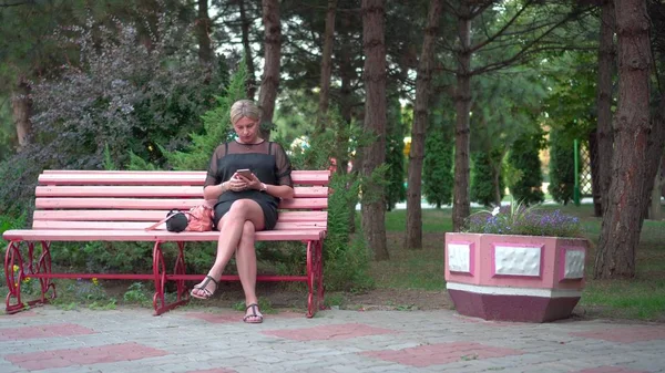 Una joven está descansando en un parque. Se sienta en un banco y mira su teléfono inteligente — Foto de Stock