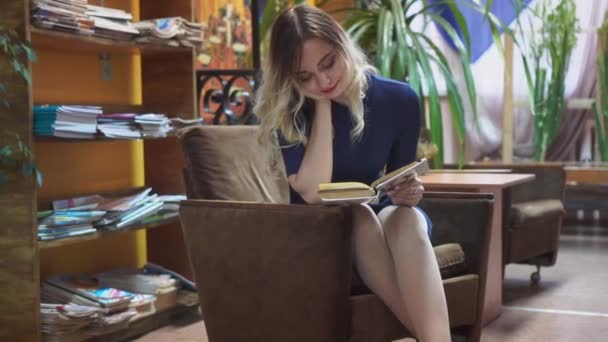 女学生在图书馆看书 一个漂亮的姑娘坐在图书馆的扶手椅上饶有兴致地看书 — 图库视频影像