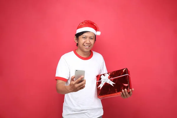 携帯電話のクリスマスアジア人男性は赤い背景で贈り物を持っている — ストック写真