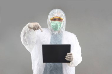 Koruyucu koruyucu koruyucu PPE giysili Asyalı doktor gri arka planda tablet gösteren tıbbi lateks eldivenler giyiyor.