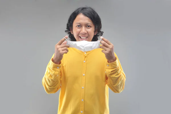 フェイスマスクを使ってアジア系の男が喜ぶ表情ですCovid 19と黄色のシャツをグレーの背景に隔離 — ストック写真