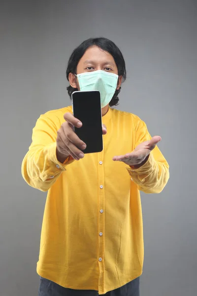 new normal asian man showing smartphone with face mask and yellow shirt isolated on gray background