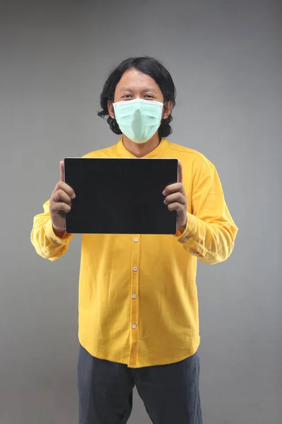 asian man new normal showing and holding tablet computer with face mask and yellow shirt isolated on gray background