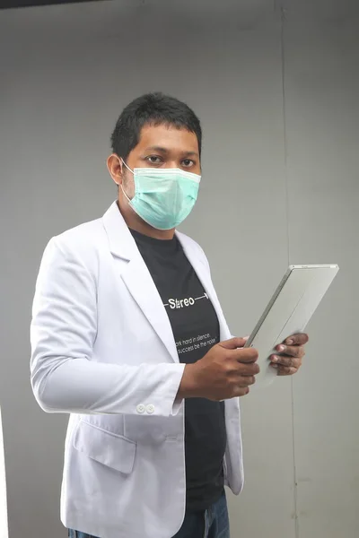 new normal singapore man doctor covid-19 using face mask holding tablets computer isolated on grey background. yogyakarta indonesia. Sep.29, 2020