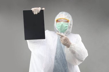 Koruyucu giysi, Google, eldiven, solunum cihazı beyaz koruyucu giysi ve maske takan doktor gri arka planda tablet gösteriyor.