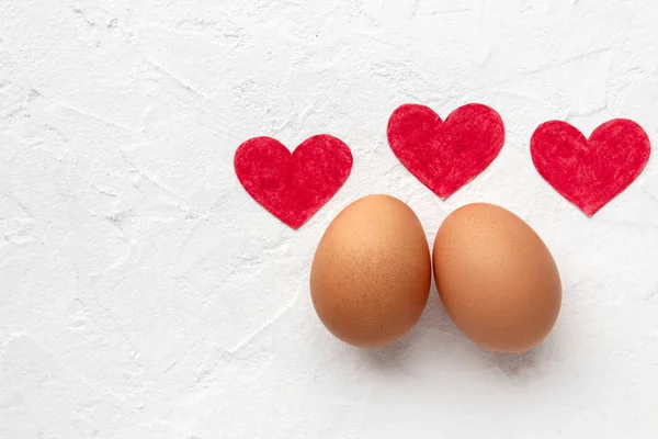 Two fresh eggs with three red paper hearts lie on white background with egg's shadow