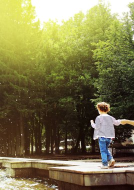 Sevimli küçük bir çocuk havuzun kenarına yürüyor, bir yetişkinin elini tutar. yeşil ağaçlar ve güneş ışığı, yetiştirme ve mutlu çocukluk kavramını arka planı