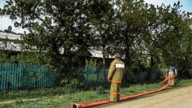 Urman, Rusya - 28 Mayıs 2011: Üniformalı iki itfaiyeci, yaz aylarında yol kenarında, rustik bir kulübe ve ahşap bir fe yangını söndürmeye hazırlanırken susuz uzun bir kırmızı yangın hortumu tutmak