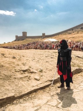 Sudak - 16 Ağustos 2015: Yıldız Savaşları destanı karakteri Darth Vader elinde bir kılıçla kale duvarının arka planında, çöl ve kalabalıklar, kostüm üretimi, siyah ceketli ve siyah maskeli bir adam, Cenova kask festivali