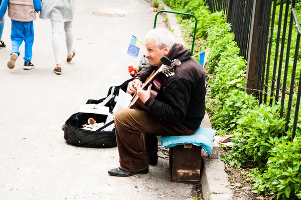 Старик-уличный музыкант играет на гитаре. Флаг Украины на заднем плане — стоковое фото