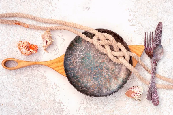 cutting board, plate, cutlery and rope sea knot with seashells on a textured concrete background, top view with copy space. sea cruise travel vacation menu trmplate.