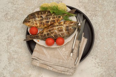 Limon ve domates ile bir tabak ve siyah daire tepsi üzerinde iki ızgara dorado balık, masa örtüsü, çatal ve bıçak ile servis, üst görünümü.