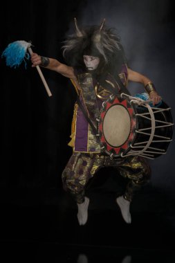 Taiko peruklu ve şeytani maskeli davulcu sahnedeki bagetlerle ve davullarla dumanlı siyah bir arka planda zıplıyor..