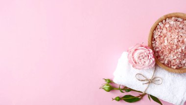 Spa yassı kompozisyonu. Ahşap kavanozda deniz tuzu, havlu, pembe arka planda çiçek. Üst görünüm, telif alanı, afiş. Günlük bakım konsepti, rahatlama ve dinlenme, banyo prosedürü.