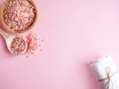 Spa yassı kompozisyonu. Tahta kavanozda deniz tuzu ve kaşıktan saçılmış, pembe arka planda banyo havlusu. Kopyalama alanı, üst görünüm. Evde bakım konsepti, rahatlama ve dinlenme, banyo prosedürü.