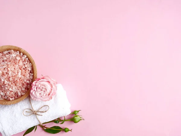 Spa yassı kompozisyonu. Ahşap kavanozda deniz tuzu, havlu, pembe arka planda çiçek. Üst görünüm, telif alanı. Günlük bakım konsepti, rahatlama ve dinlenme, banyo prosedürü.
