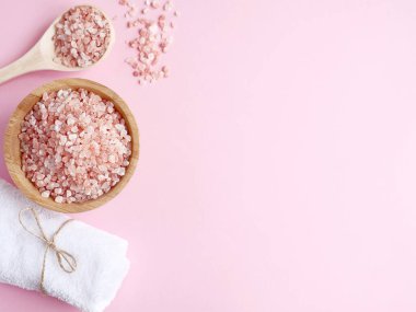 Spa yassı kompozisyonu. Tahta kavanozda deniz tuzu ve kaşıktan saçılmış, pembe arka planda banyo havlusu. Kopyalama alanı, üst görünüm. Evde bakım konsepti, rahatlama ve dinlenme, banyo prosedürü.