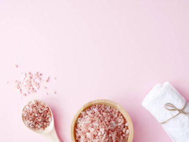 Spa yassı kompozisyonu. Tahta kavanozda deniz tuzu ve kaşıktan saçılmış, pembe arka planda banyo havlusu. Kopyalama alanı, üst görünüm. Evde bakım konsepti, rahatlama ve dinlenme, banyo prosedürü.
