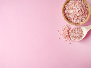Spa yassı kompozisyonu. Tahta kavanozda deniz tuzu ve kaşıktan saçılmış, pembe arka planda banyo havlusu. Kopyalama alanı, üst görünüm. Evde bakım konsepti, rahatlama ve dinlenme, banyo prosedürü.