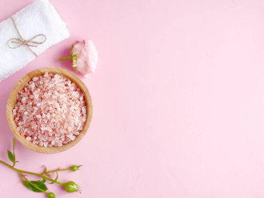 Spa yassı kompozisyonu. Ahşap kavanozda deniz tuzu, havlu, pembe arka planda çiçek. Üst görünüm, telif alanı. Günlük bakım konsepti, rahatlama ve dinlenme, banyo prosedürü.