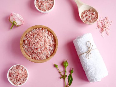 Spa yassı kompozisyonu. Tahta kavanozda deniz tuzu ve kaşıktan, havludan, çiçekten pembe arka planda serpilmiş. Üst Manzara. Günlük bakım konsepti, rahatlama ve dinlenme, banyo prosedürü