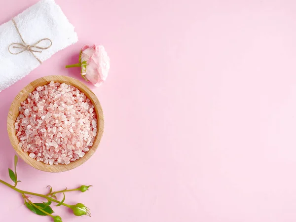 Spa yassı kompozisyonu. Ahşap kavanozda deniz tuzu, havlu, pembe arka planda çiçek. Üst görünüm, telif alanı. Günlük bakım konsepti, rahatlama ve dinlenme, banyo prosedürü.