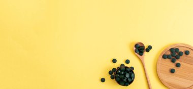 Sarı arka planda tahta kaşık ve kavanozla tabletlerde spirulina. Doğal, eko, toz sağlıklı beslenme takviyesi kavramı. Kopyalama alanı, pankart.