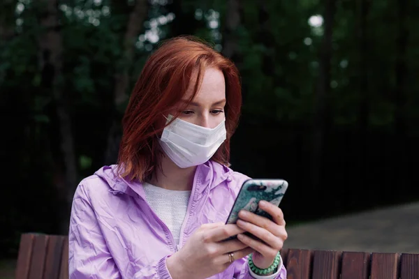 Coronavirus canlandırması sırasında parktaki akıllı telefondan haberleri okuyan maskeli kızıl saçlı kız.