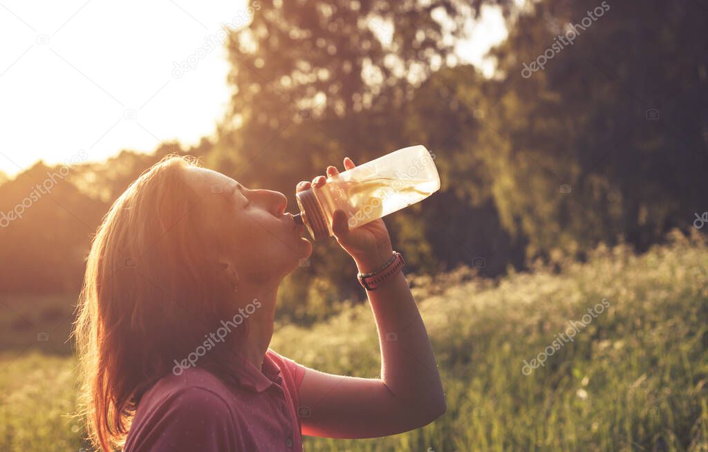 Girl greedily drinking water with lemons from clear plastic bottle in contrast light during summer sunset