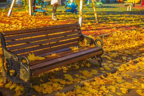 Çocuk parkının yanındaki bankta sarı yapraklar ve seçici bir odak noktası var.