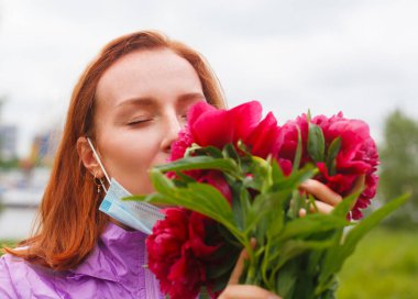 Revival after coronavirus. Redhead girl sniffs pink flowers with pulled down medicine mask. Recovery after coronavirus and flu. Sense of smell return. Discharge from hospital. clipart