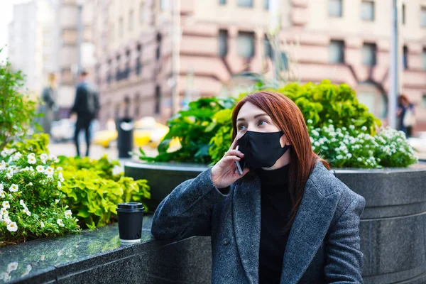 Redhead woman in protective mask talking on a smartphone in downtown. Business during coronavirus concept. Woman in black mask calling on a phone close-up