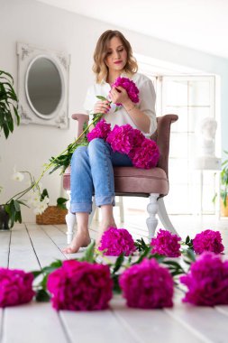 Beyaz gömlekli ve kot pantolonlu bir kadın ellerinde pembe şakayıklar, beyaz bir odada bir sandalyede oturuyor..