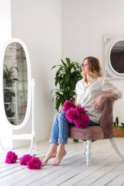 Beyaz gömlekli ve kot pantolonlu bir kadın ellerinde pembe şakayıklar, beyaz bir odada bir sandalyede oturuyor..