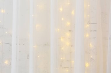 Yeni yıl ya da Noel geçmişi. Tebrik kartı. Beyaz tül perde arka planda, yanan bir çelenkle dekore edilmiş beyaz tuğla bir duvar..