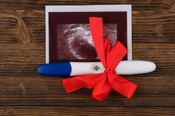 Photo of ultrasound and positive results pregnancy test with red bow on wooden backgrounds.