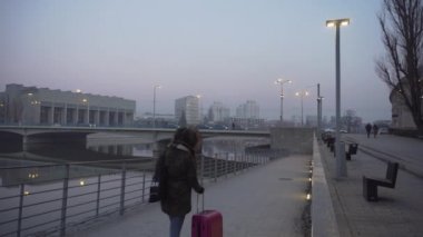 Pembe bavullu kız Wroclaw'da sisli bir nehir kıyısında yürüyor.