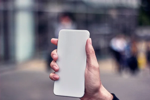 Bulanık bir şehrin arka planında logosu olmayan beyaz bir telefon tutan genç bir adam.
