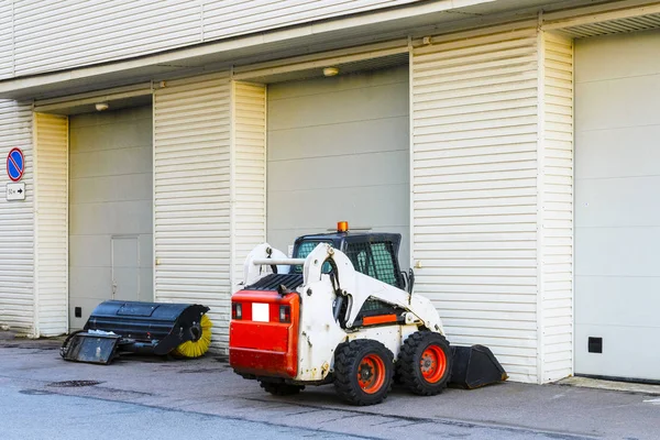 a small mini tractor with a blade and a bucket for street cleaning is at the gate of a large garage