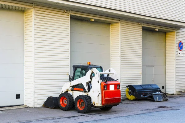 a small mini tractor with a blade and a bucket for street cleaning is at the gate of a large garage