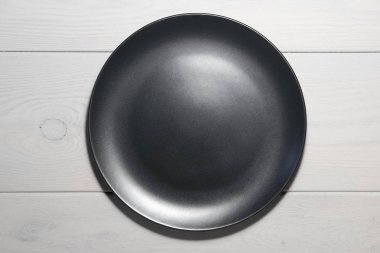 Siyah yuvarlak boş bir tabak, ahşap desenli, hafif gri bir ahşap zemin üzerinde duruyor. Yukarıdan görünüm.