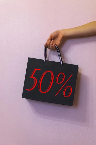 black gift shopping bag with 50% sale, holds in hand on pink background.