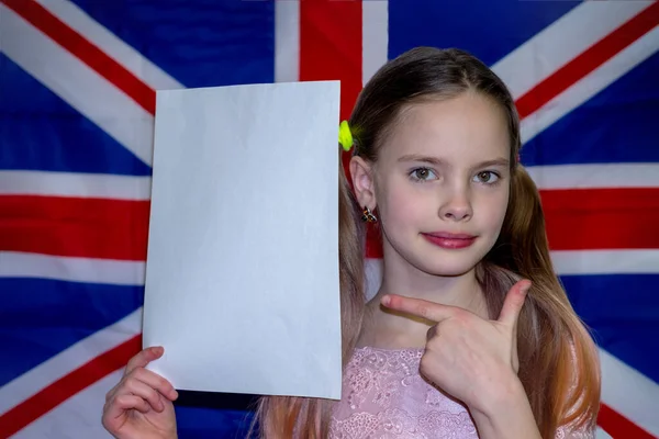 A girl with blond hair and two pigtails on the background of the flag of Great Britain holds a white sheet of paper. background in the form of an English flag with a beautiful, emotional, smiling girl